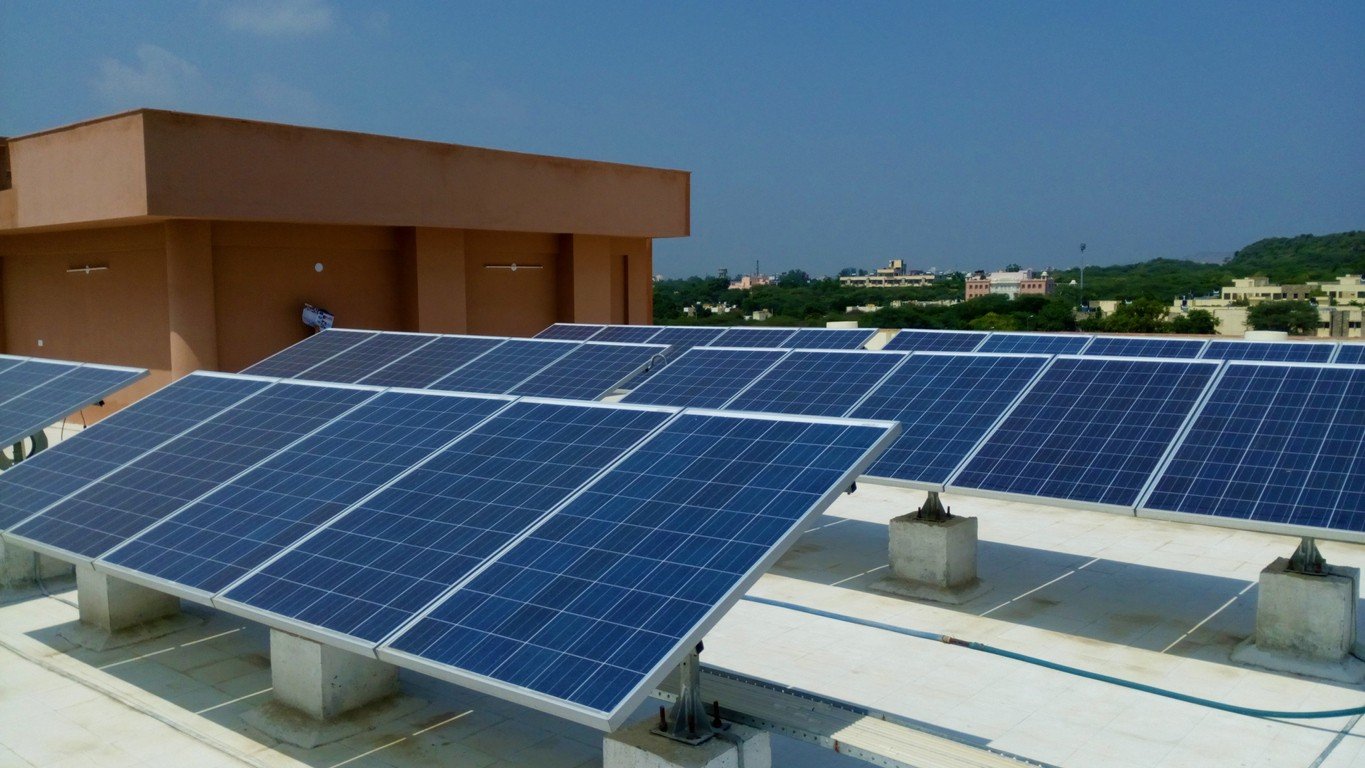 Three rows of solar panels on roof of a California college building with part of the building and other buildings in the background.